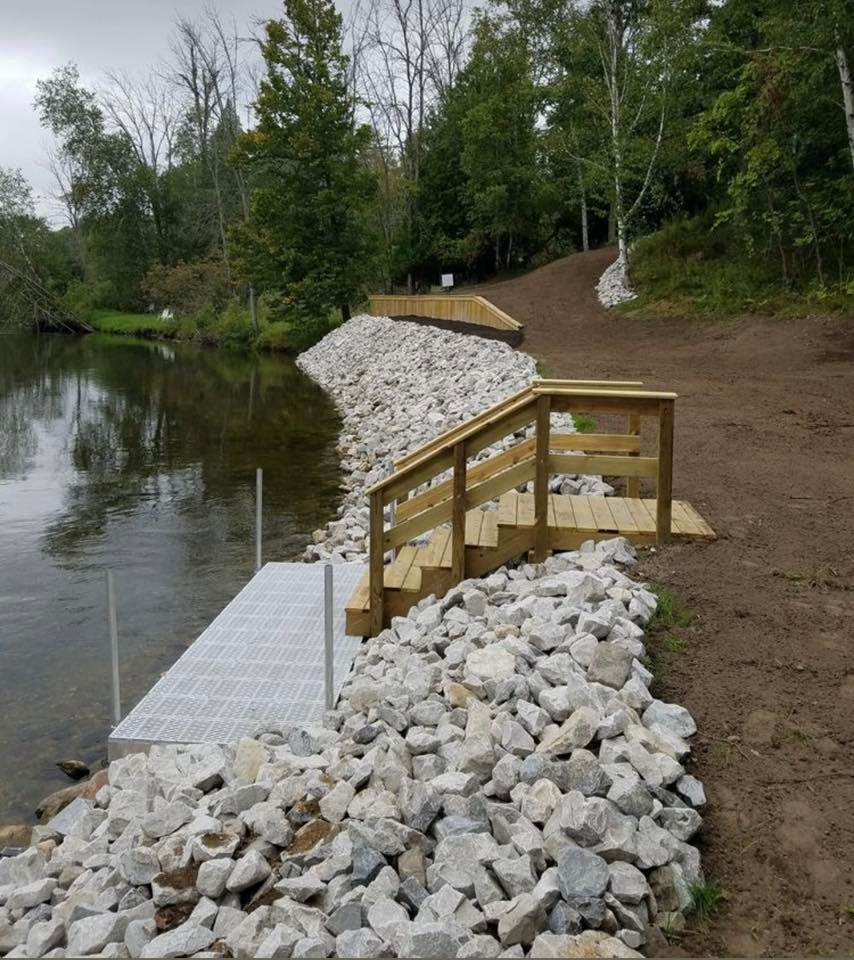 Wooden ramp and stairs beside a lake, bordered by rocks and trees on a dirt path.