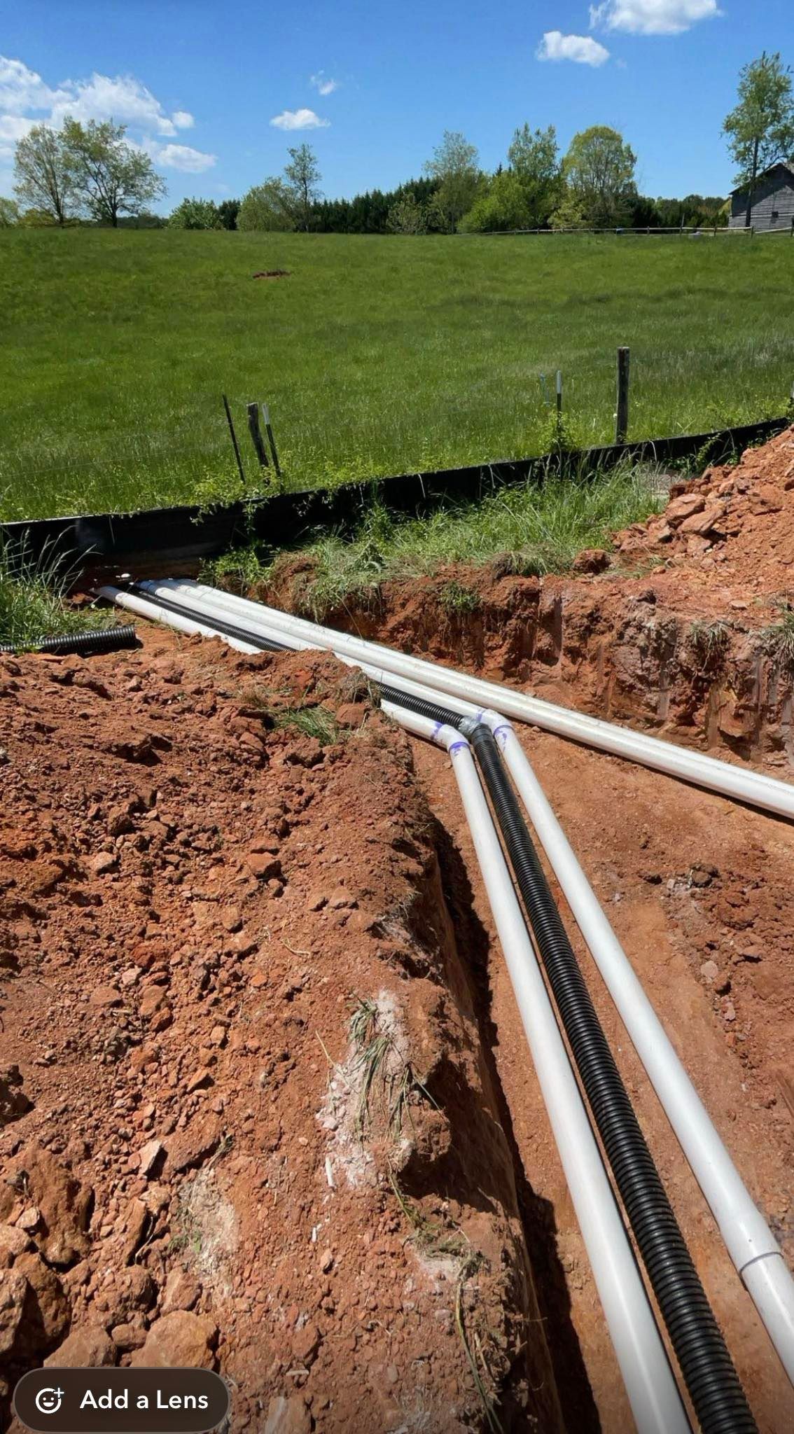Trenches with white utility pipes laid across red soil beside a grassy field under a blue sky