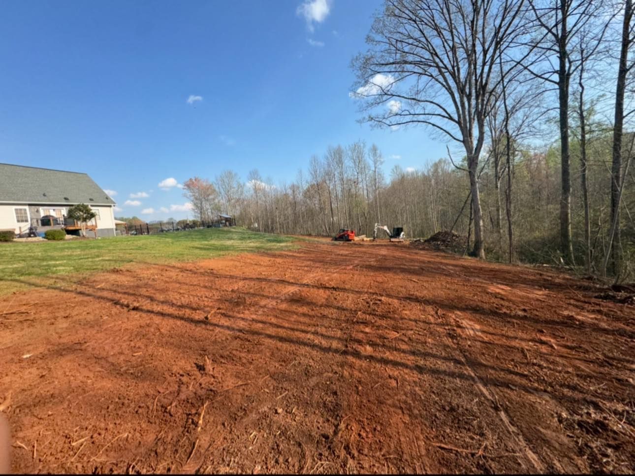 Red dirt lot beside a house and trees under a clear blue sky