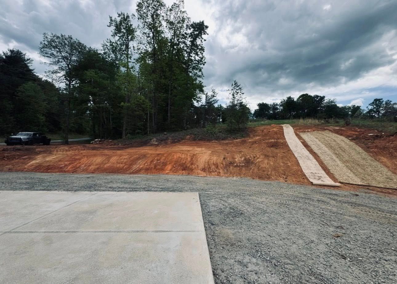 Gravel driveway and red dirt slope beside a concrete slab under cloudy sky, with trees in the background