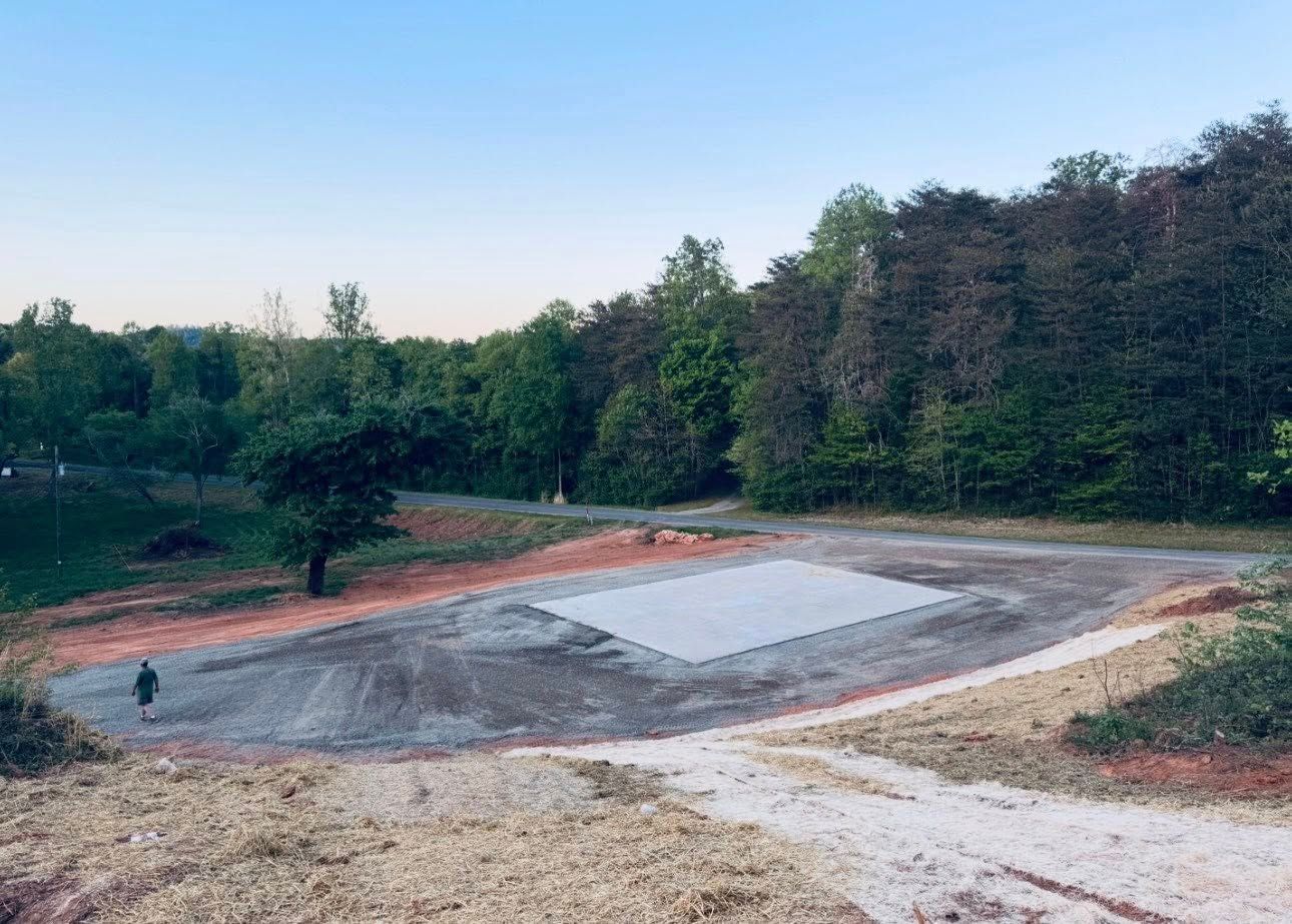 Construction site with a cleared dirt area, gravel pad, and trees surrounding the site at dusk
