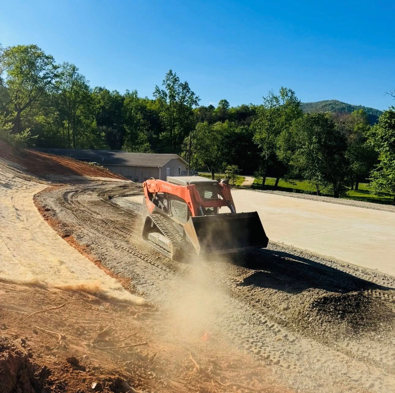 Orange skid steer spreading gravel on a dirt road beside trees and a blue sky