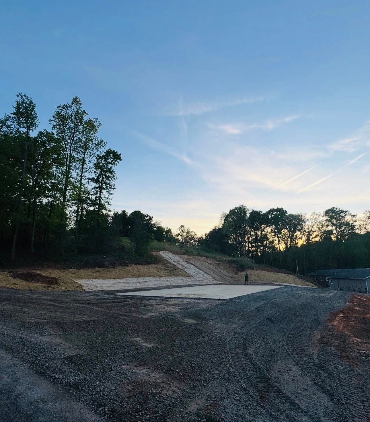 Gravel road and cleared dirt lot at dusk, bordered by trees under a clear blue sky.