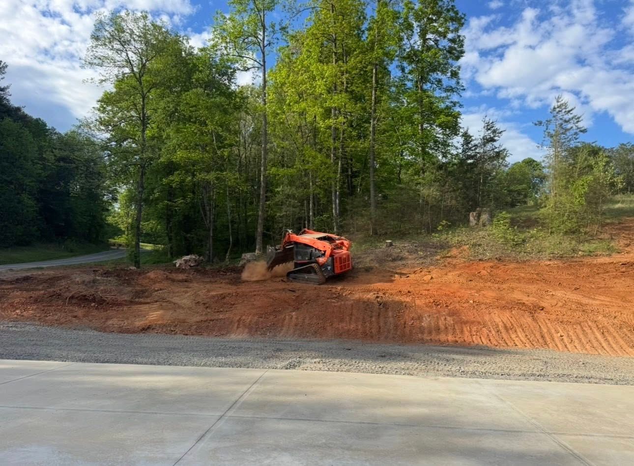 Orange excavator clearing red dirt beside a paved road in a wooded area under a blue sky