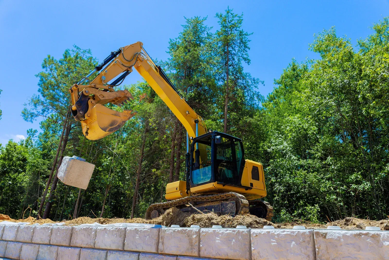 Yellow excavator lifting a concrete block above a stone wall with green trees in the background