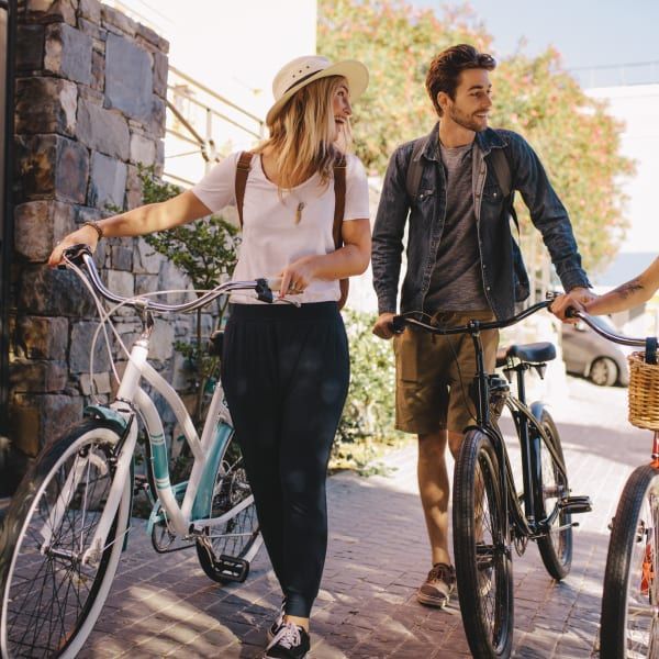 A couple walks outdoors on a sunny day, laughing while pushing their bicycles along a cobblestone path.