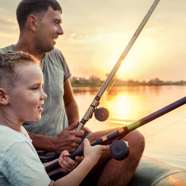 A person and a child fishing on a boat at sunset, holding fishing rods while looking toward the water.