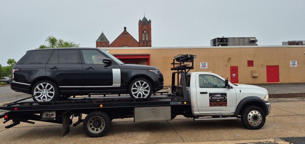 Black Range Rover SUV on a tow truck, driving past a tan building with red doors, and a church.