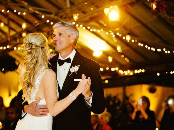 A bride and a man in a tuxedo dancing in a warmly lit, string-light-decorated venue.