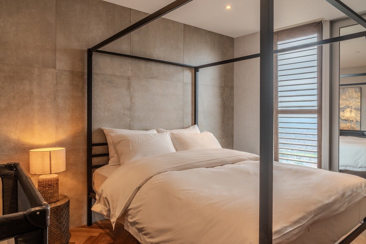 Bedroom with a four-poster bed and a window with blinds. Beige bedding and a bedside lamp.