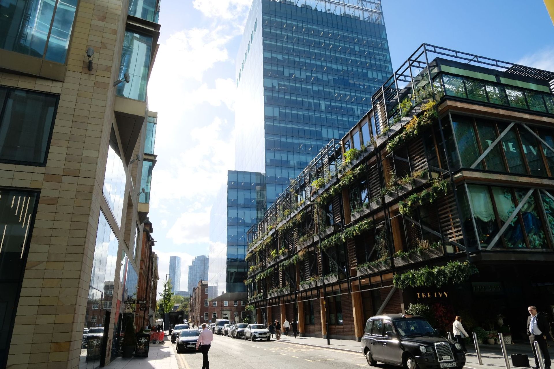 Street view of buildings in a city. Tall glass skyscraper in the background. Lower buildings with plants on balconies.