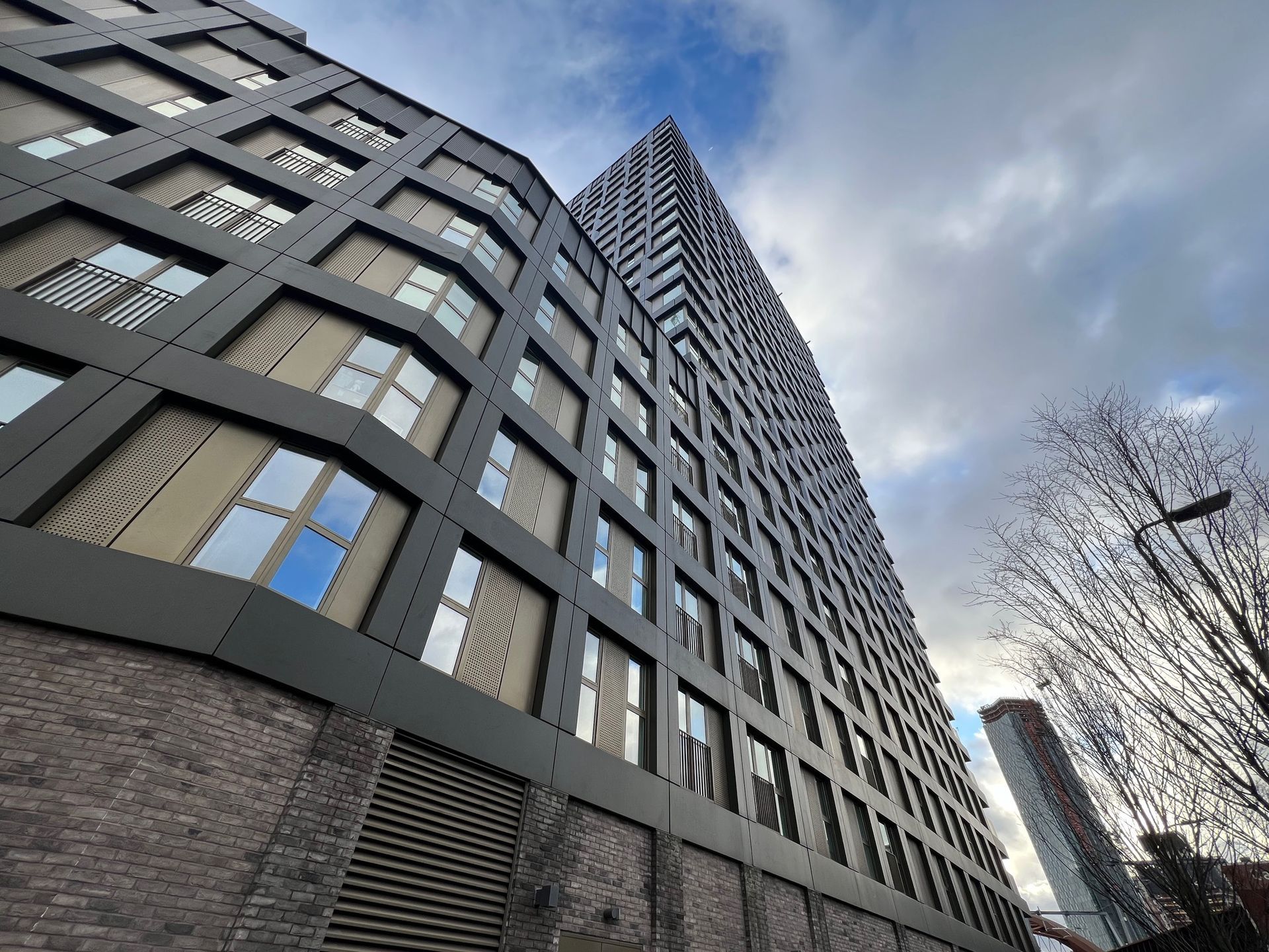 Tall modern building with dark facade, blue sky visible in windows, against a cloudy sky.