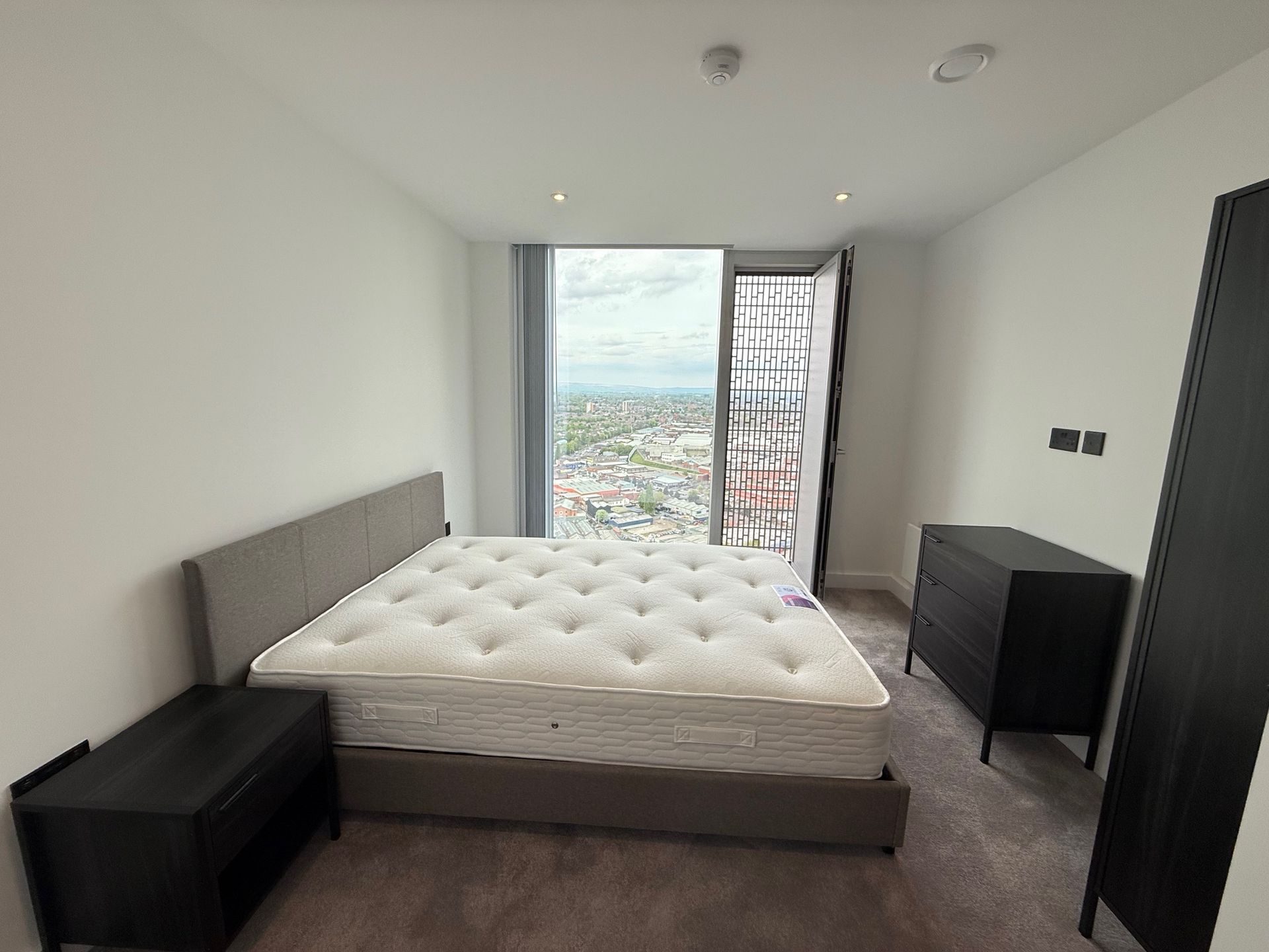 Bedroom with a bed, dresser, nightstand, and large window overlooking a city.