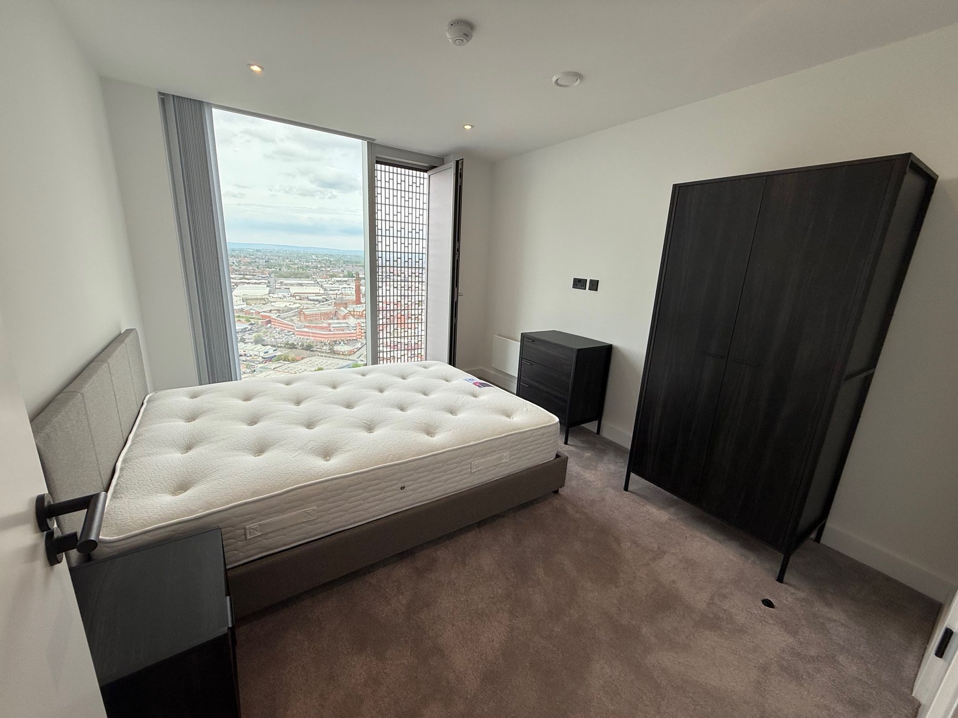 Bedroom with a bed, a wardrobe, a bedside table, and a large window overlooking the city.