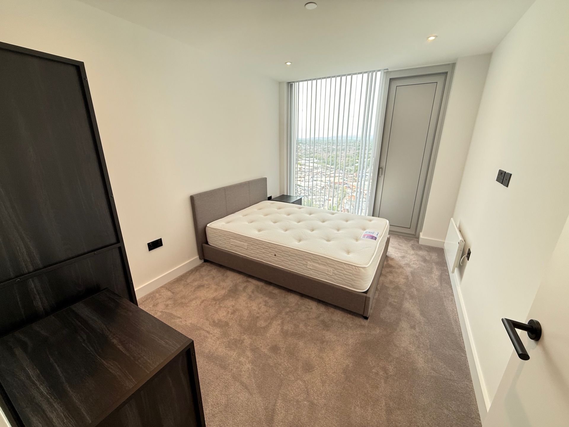 Bedroom with a bed, large window, and dark wood furniture on a carpeted floor.