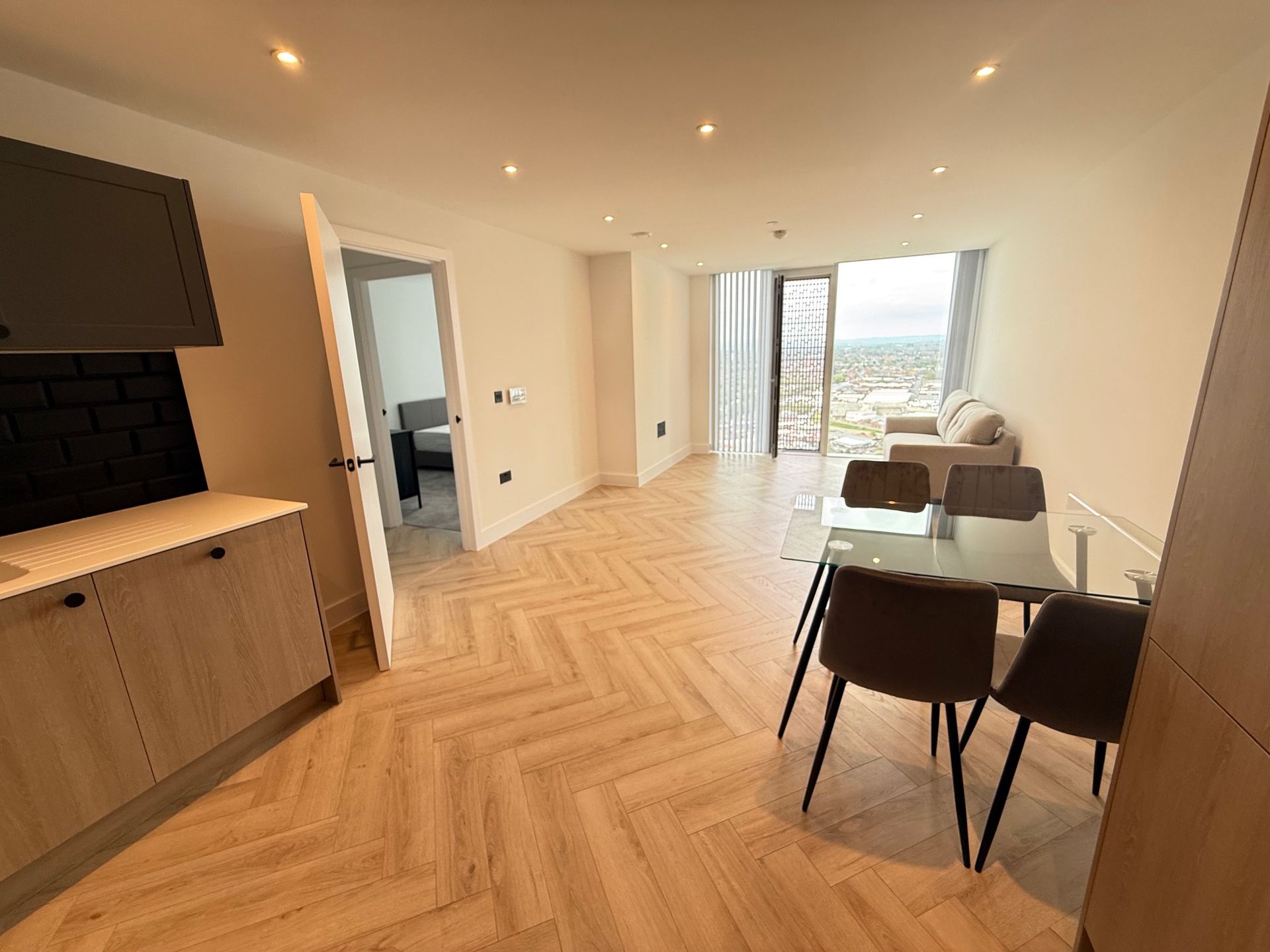 Modern apartment interior with light wood floors, kitchen, dining area, and view of city through a large window.