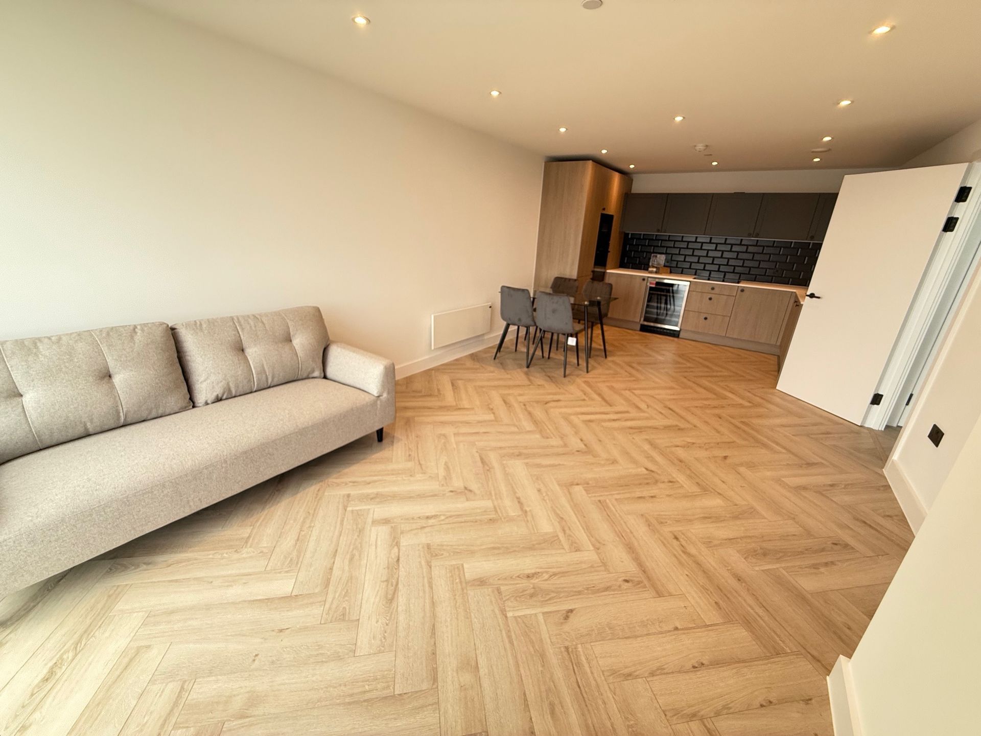 Spacious open-plan living area with herringbone wood floor, sofa, dining table, and kitchen in the background.