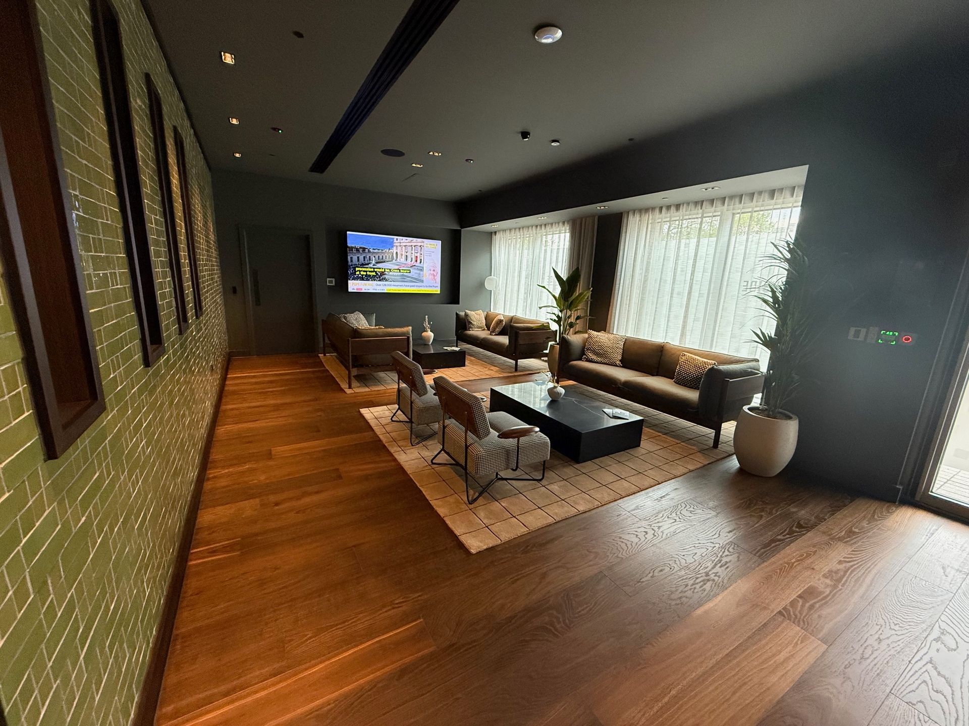 Lounge area with brown wood floors, green tile wall, and seating around a rug. TV on the wall.