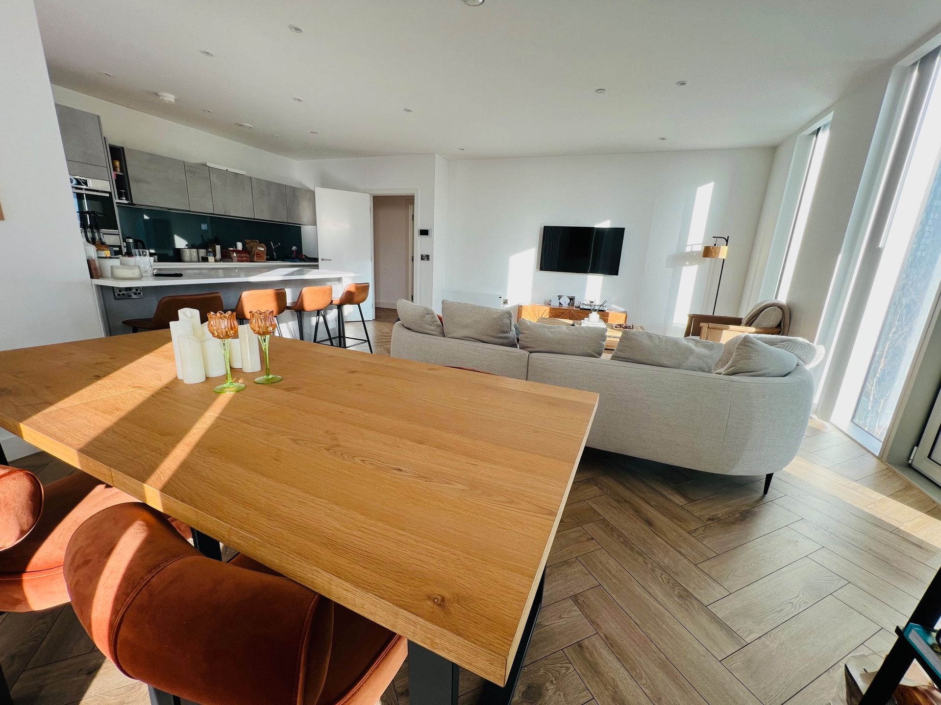 Bright living area with wooden floors, sofa, table, kitchen, and window.