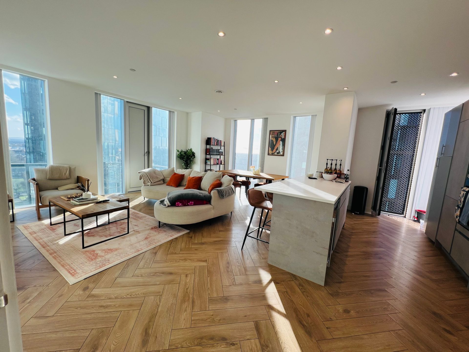 Spacious, modern living room with curved sofa, wooden floors, large windows, and a kitchen island.