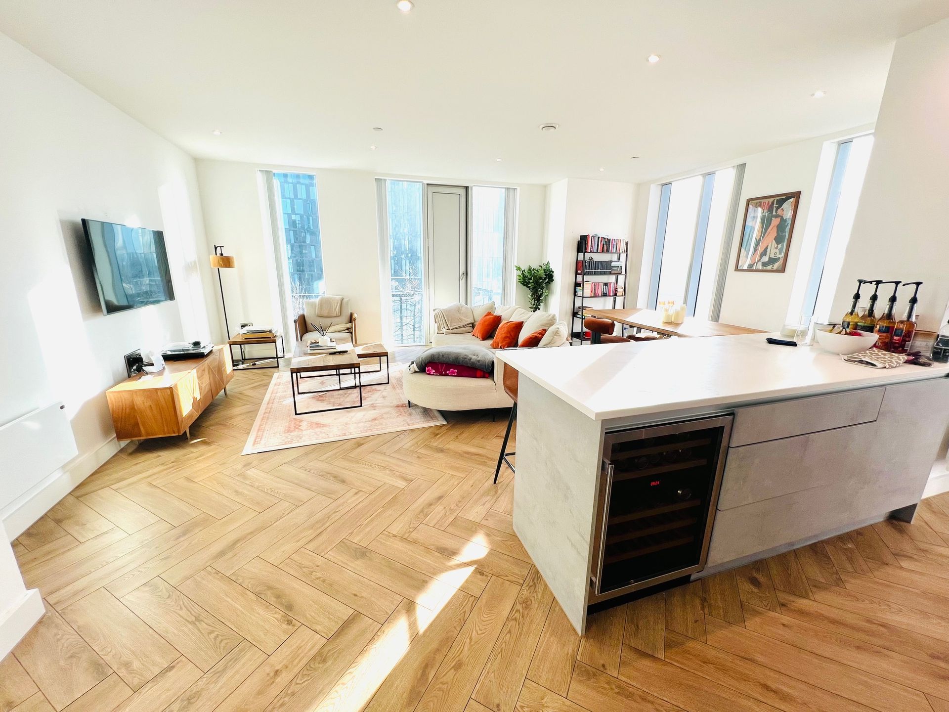 Bright living room with a kitchen island. Wooden floors, tan sofa, and large windows.