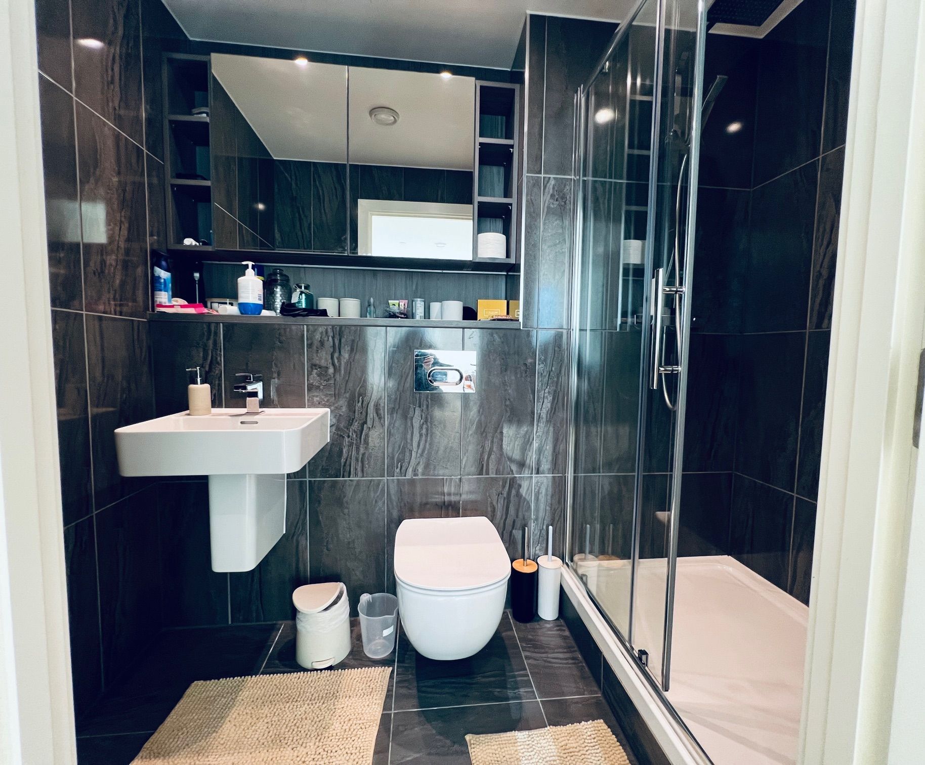 Modern bathroom with dark tile, floating sink, toilet, shower, and a large mirror.