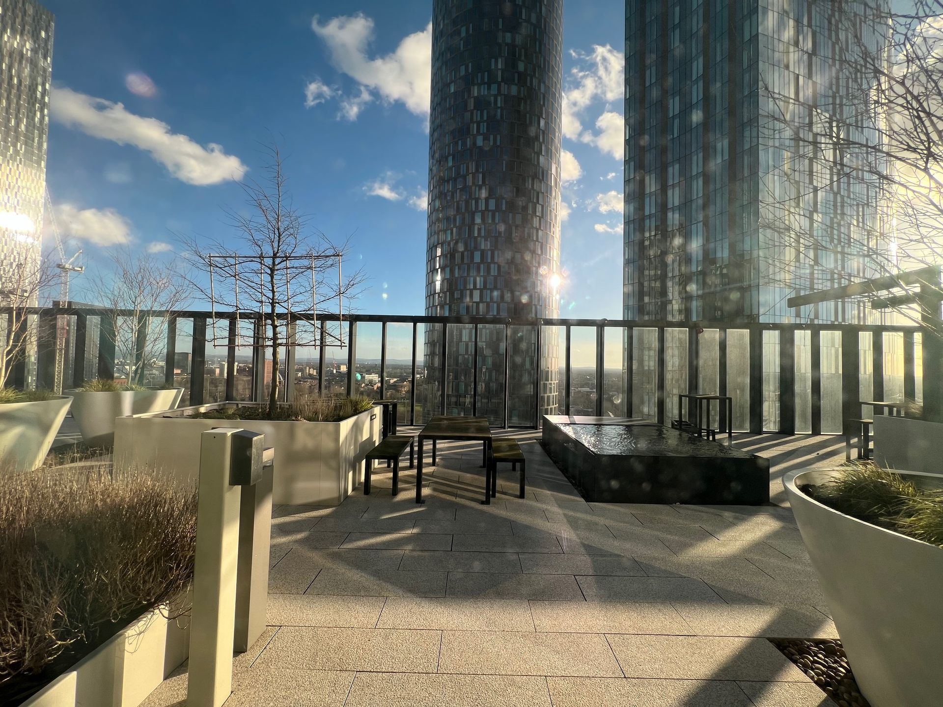 Rooftop patio with seating, planters, and tall skyscrapers in the background on a sunny day.