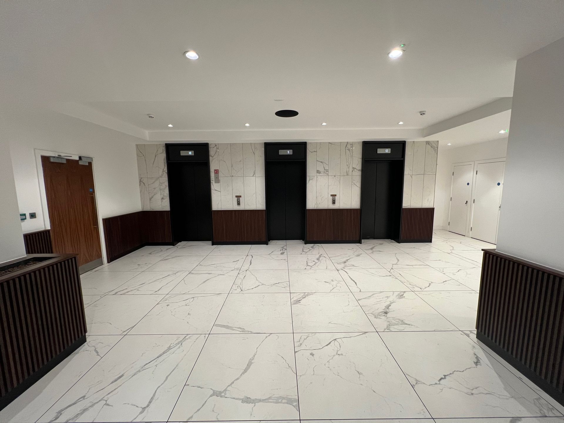Lobby with three black elevators, white marble floor, and wood paneling.