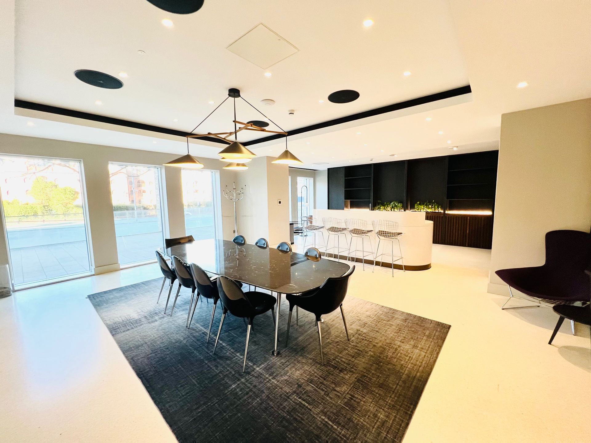 Modern dining area with long table, chairs, rug, and pendant lights. Large windows, bar and seating.