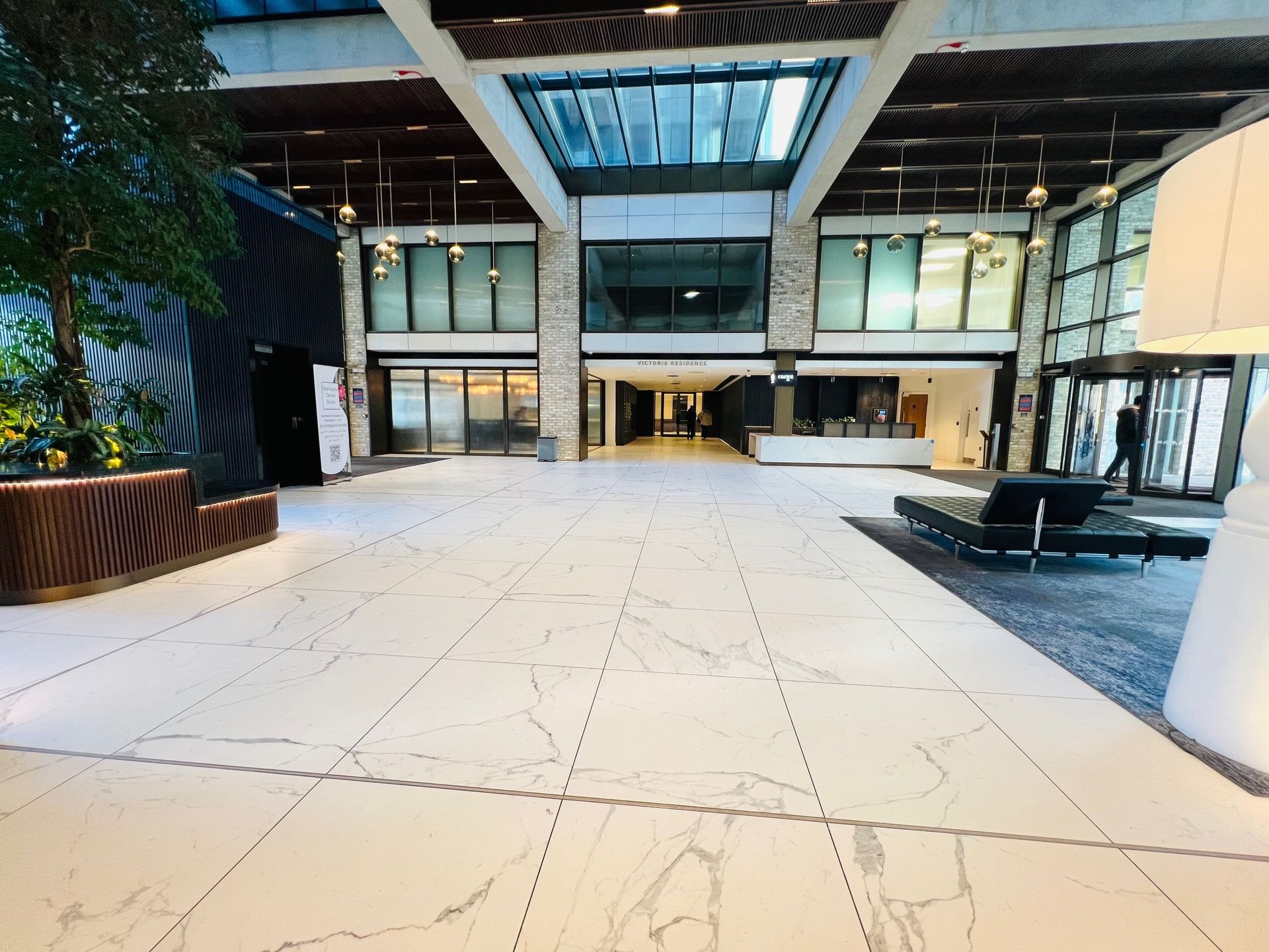 Spacious lobby with white tiled floor, glass windows, and a skylight. A person walks near the entrance.