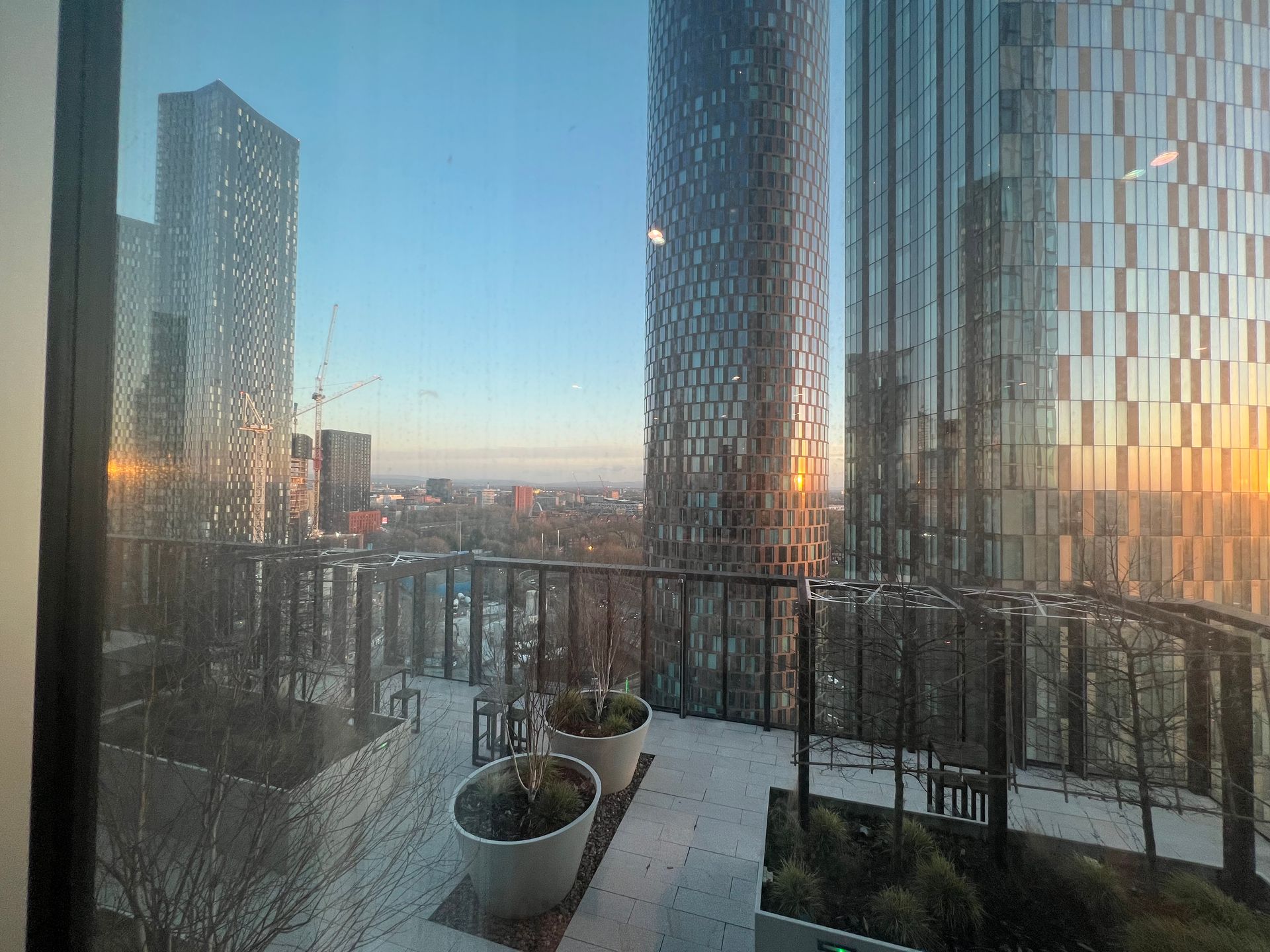 Balcony view of tall city buildings at sunset. Potted plants and metal railings are visible.
