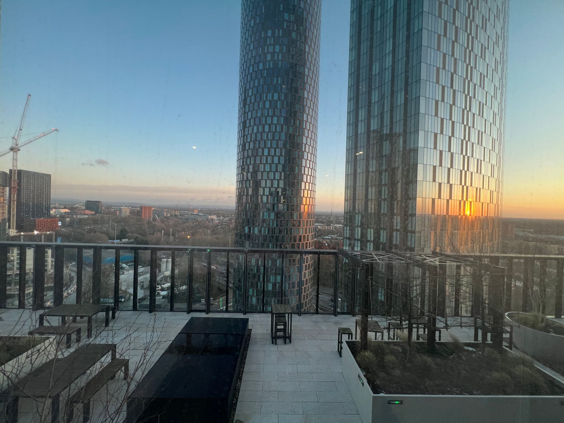 Rooftop view with two tall skyscrapers at sunset. Outdoor patio with benches, planters, and a cityscape.