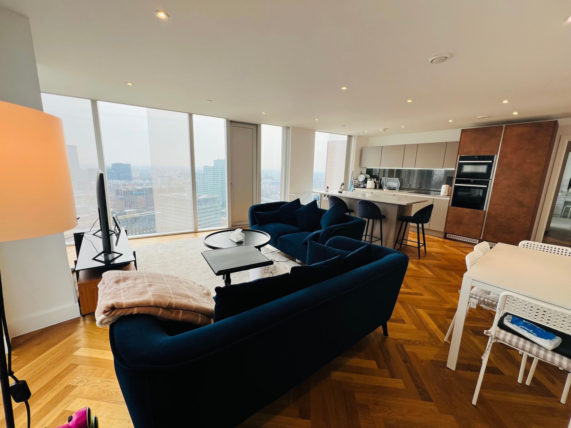 Modern living room with blue sofa, open kitchen, and city view through large windows.