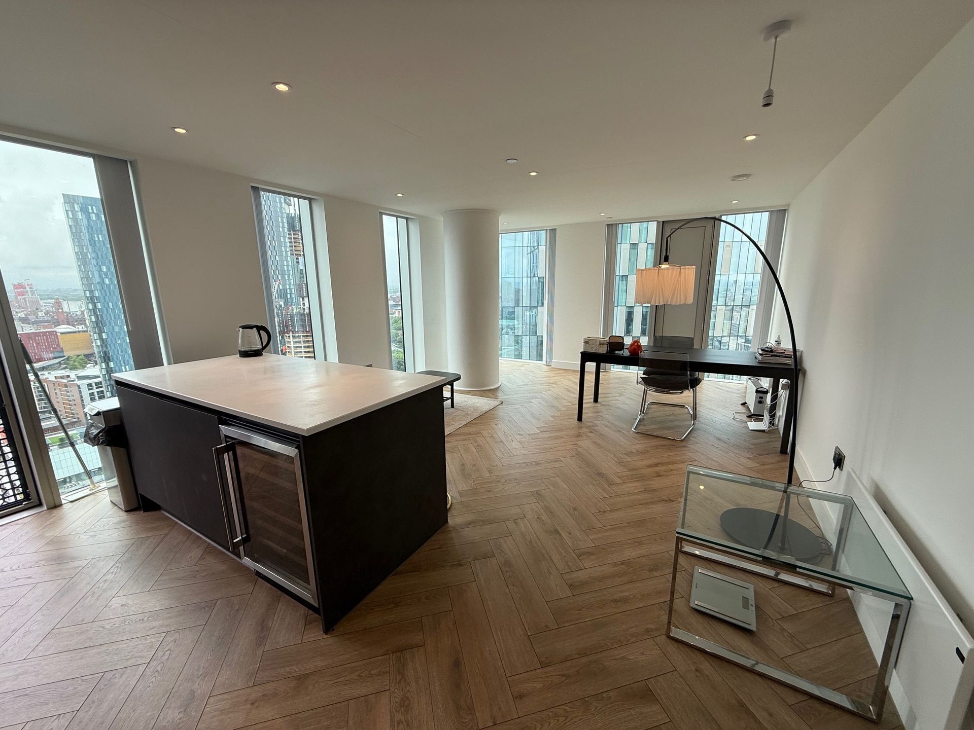 Modern kitchen/living space with island, desk, and large windows with city views. Light wood floors.