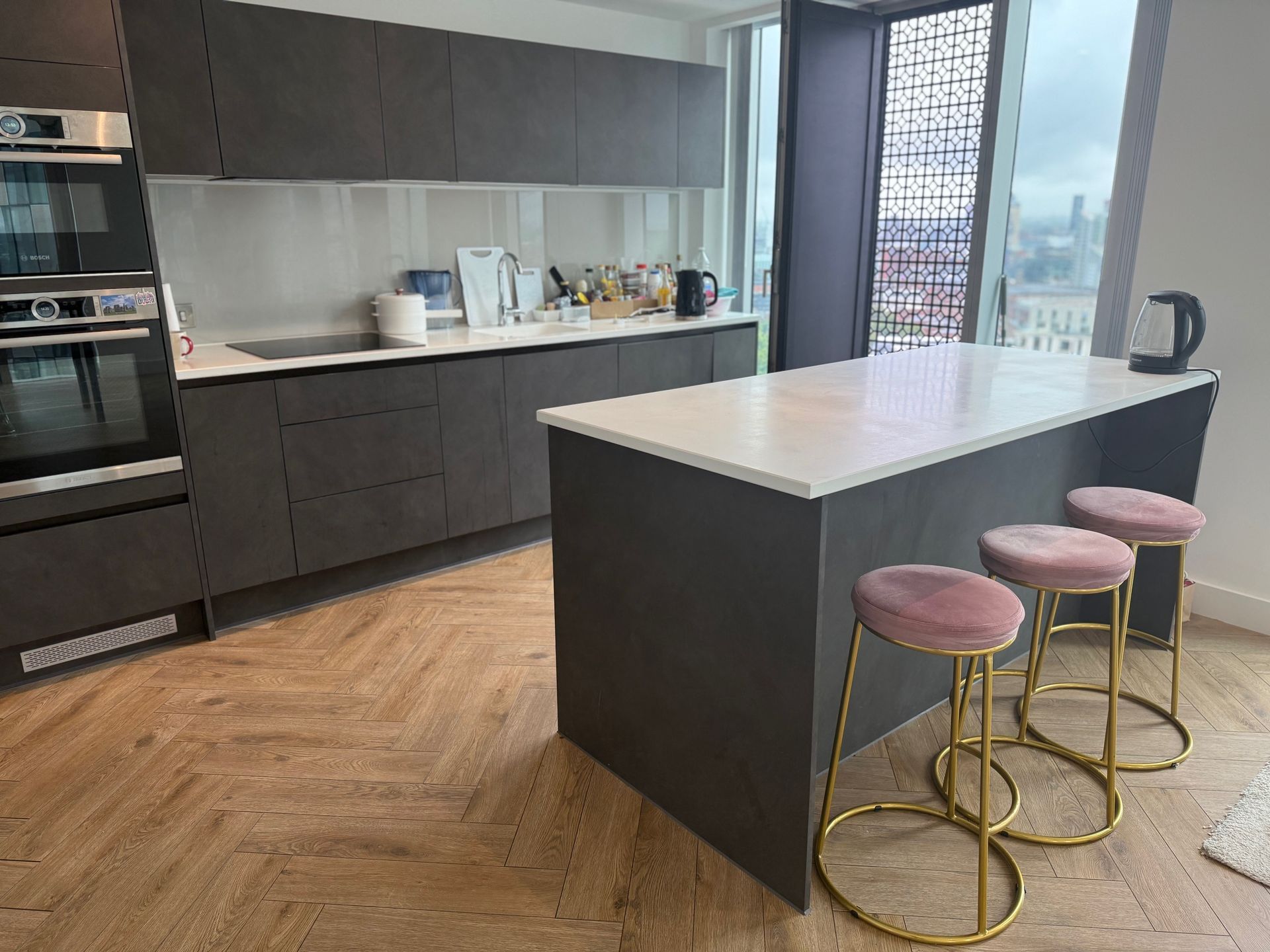 Modern kitchen with dark gray cabinets, white countertop island, pink stools, and hardwood floor.