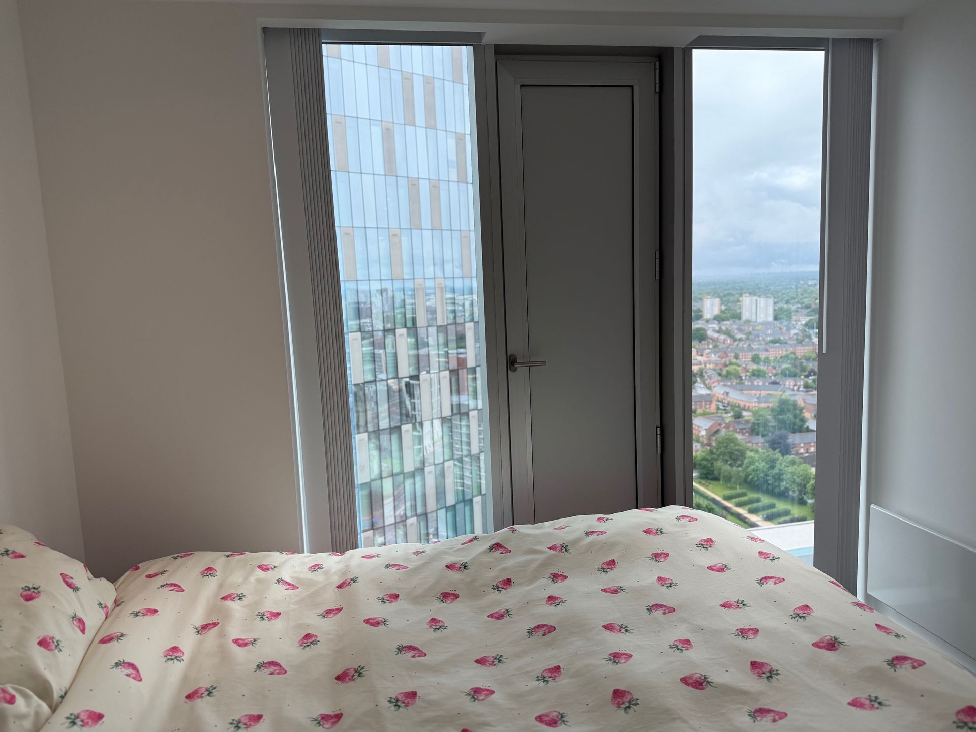 Bedroom with large windows showing cityscape and bed with floral bedding.