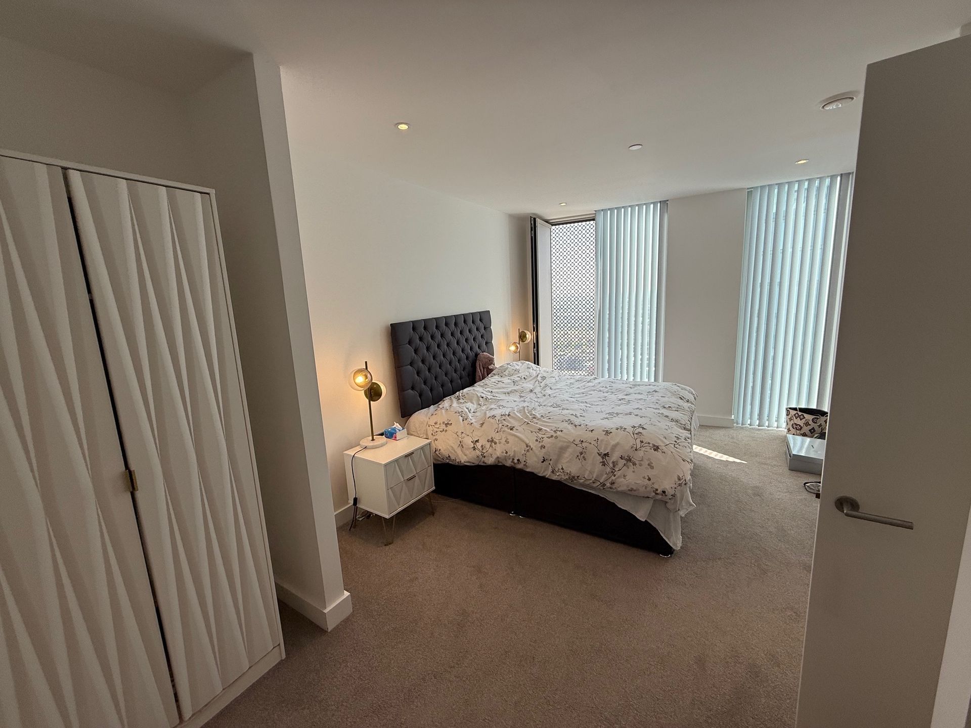 Bedroom with bed, nightstands, and textured closet door. White walls, carpet, and windows with curtains.