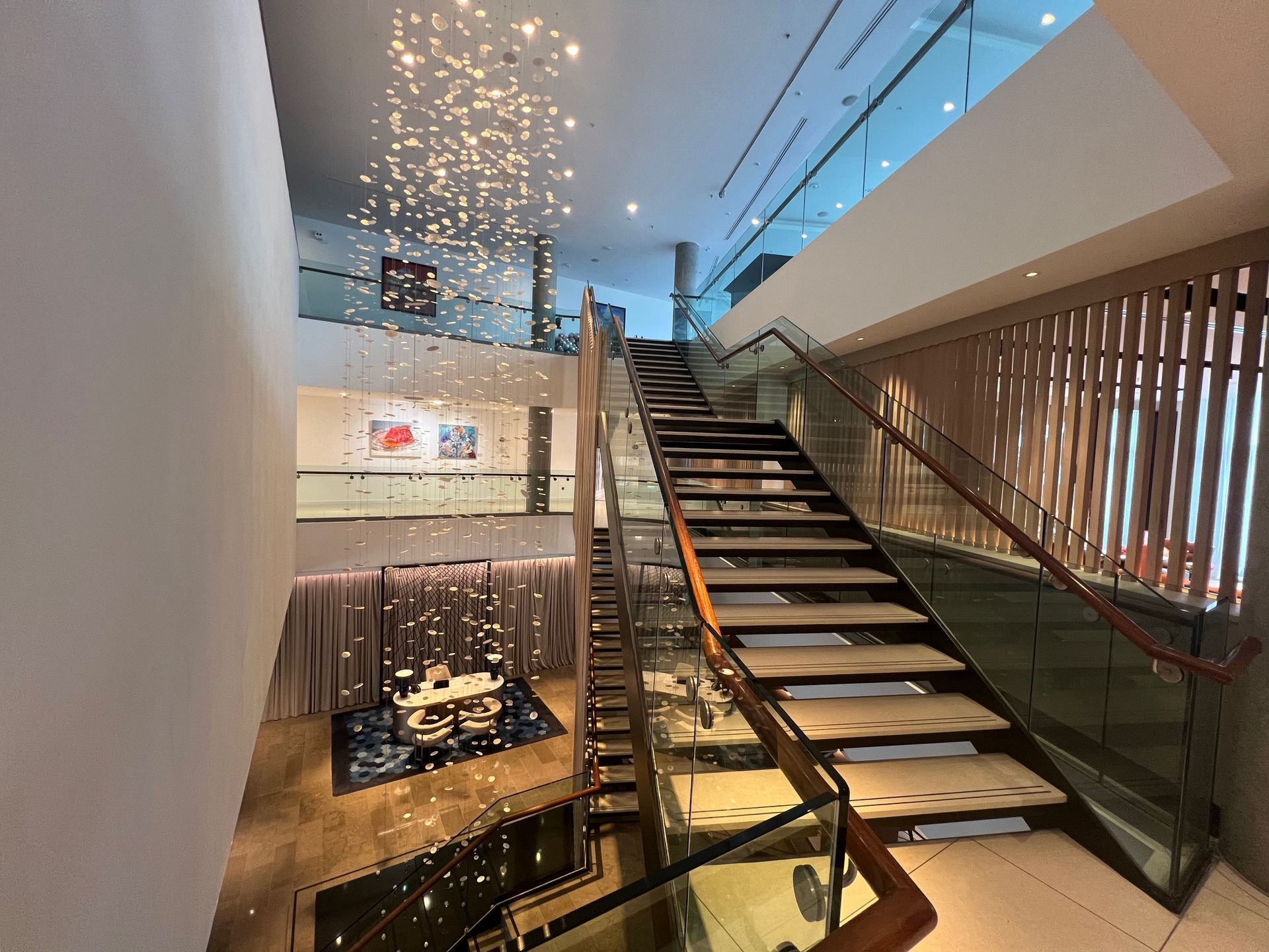 A modern staircase with glass railings, leading up to a higher level. A chandelier hangs above, illuminating the space.