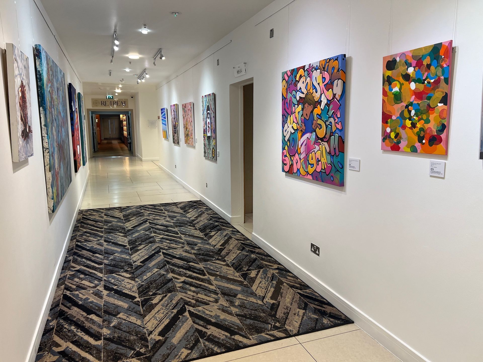 Art gallery hallway with paintings on white walls and a patterned rug.