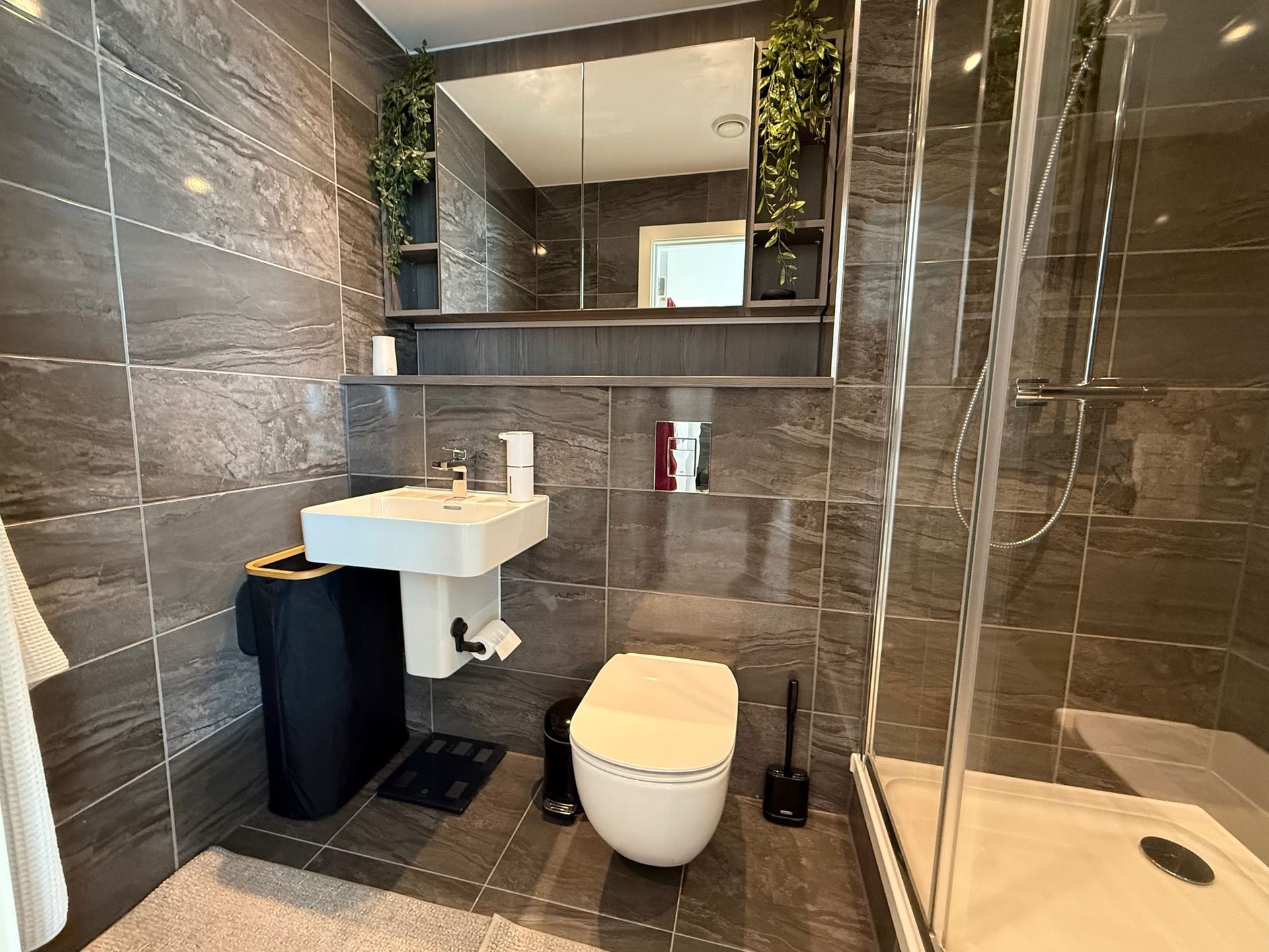Bathroom with gray tile walls and floor. Features include a sink, toilet, shower, and mirror cabinet with plants.
