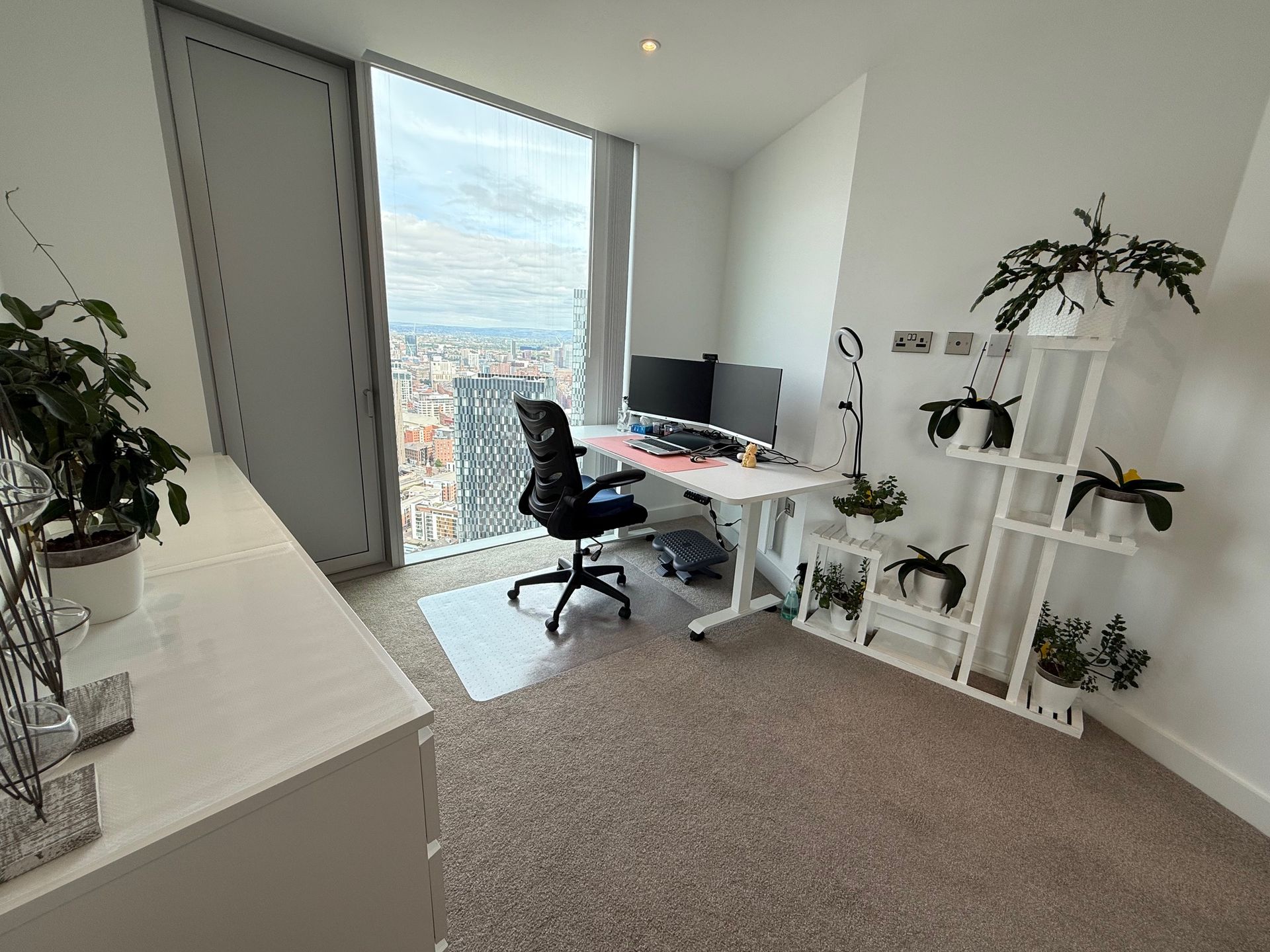 Home office with desk, monitors, chair, plants, and large window overlooking a city.
