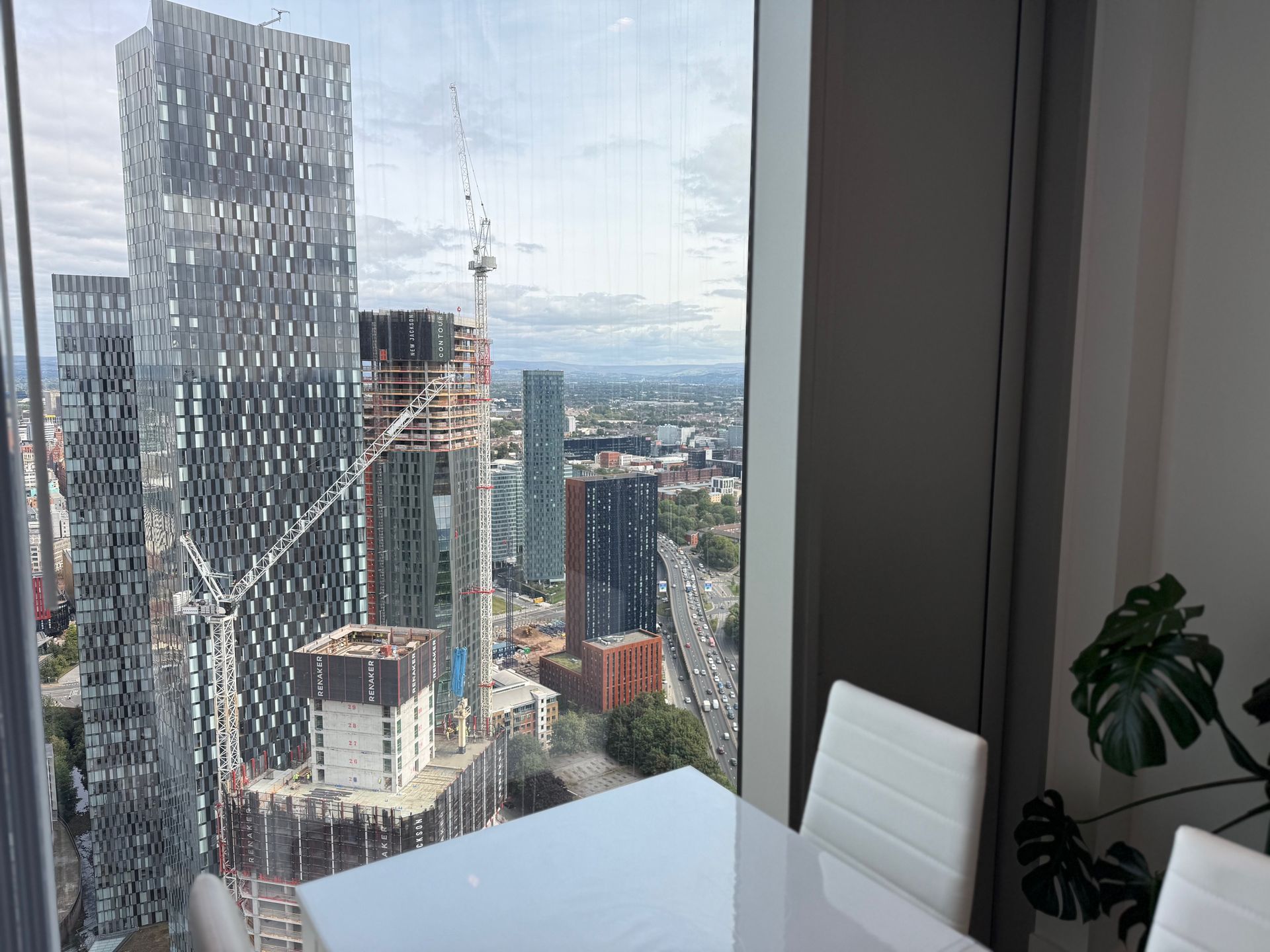 Cityscape view from a high-rise window, showing tall buildings, construction cranes, and a table with white chairs.