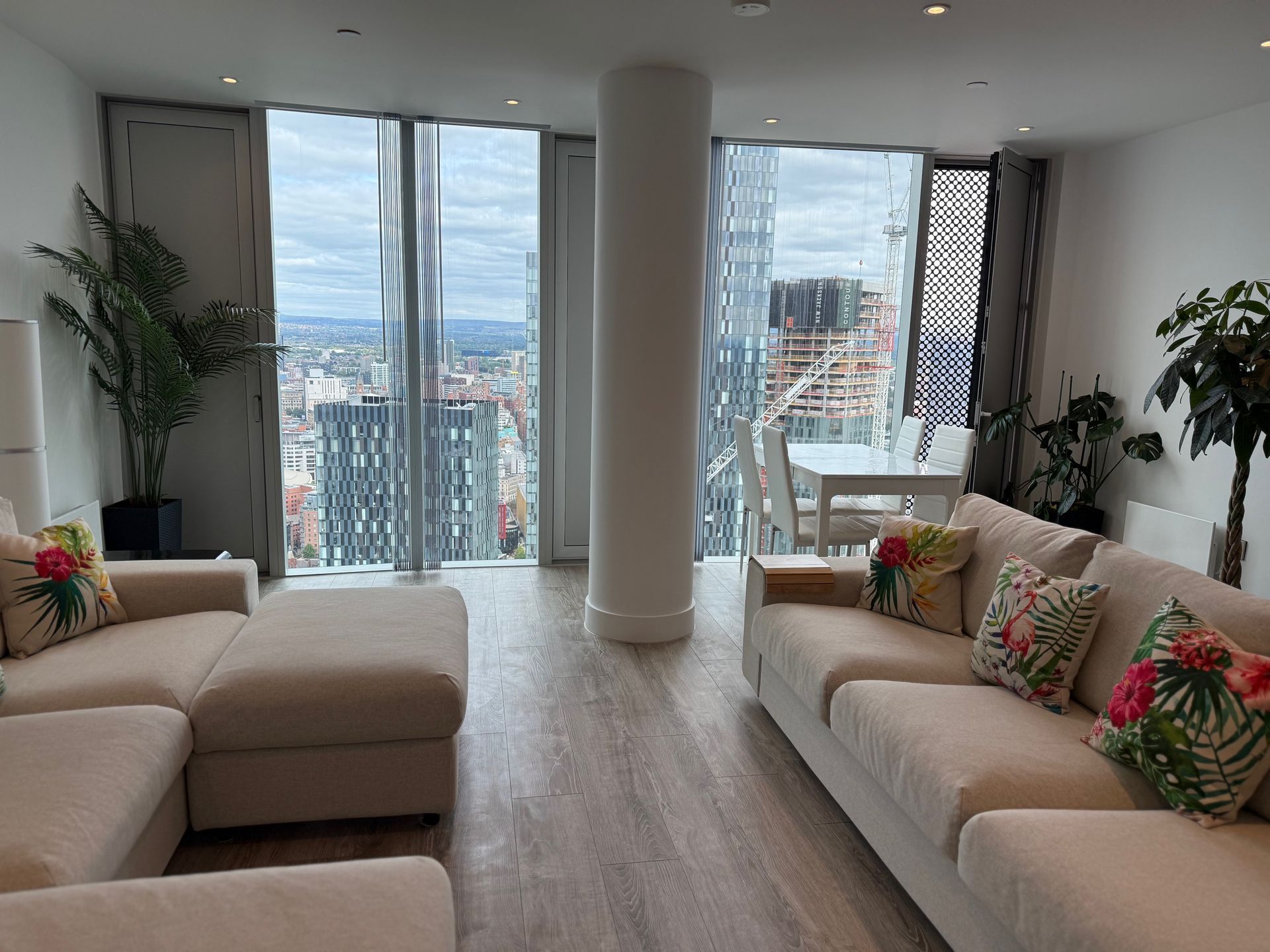 Living room with light-colored sofas, large windows showing city view, and decorative pillows.