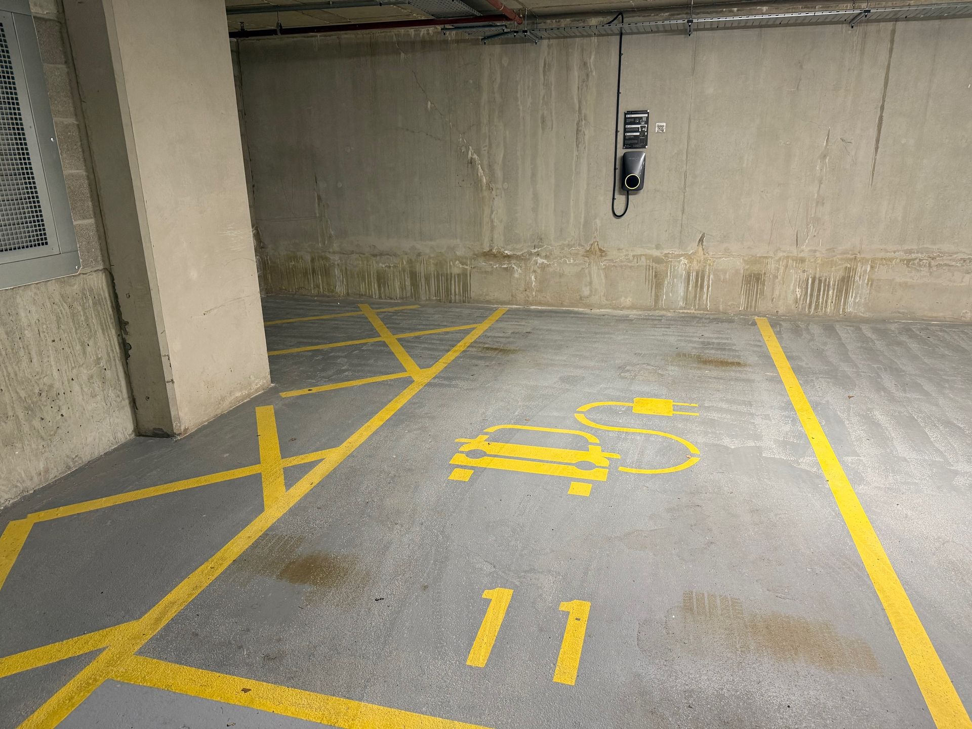 Electric vehicle charging station space in a concrete garage, marked with yellow lines and a car symbol.