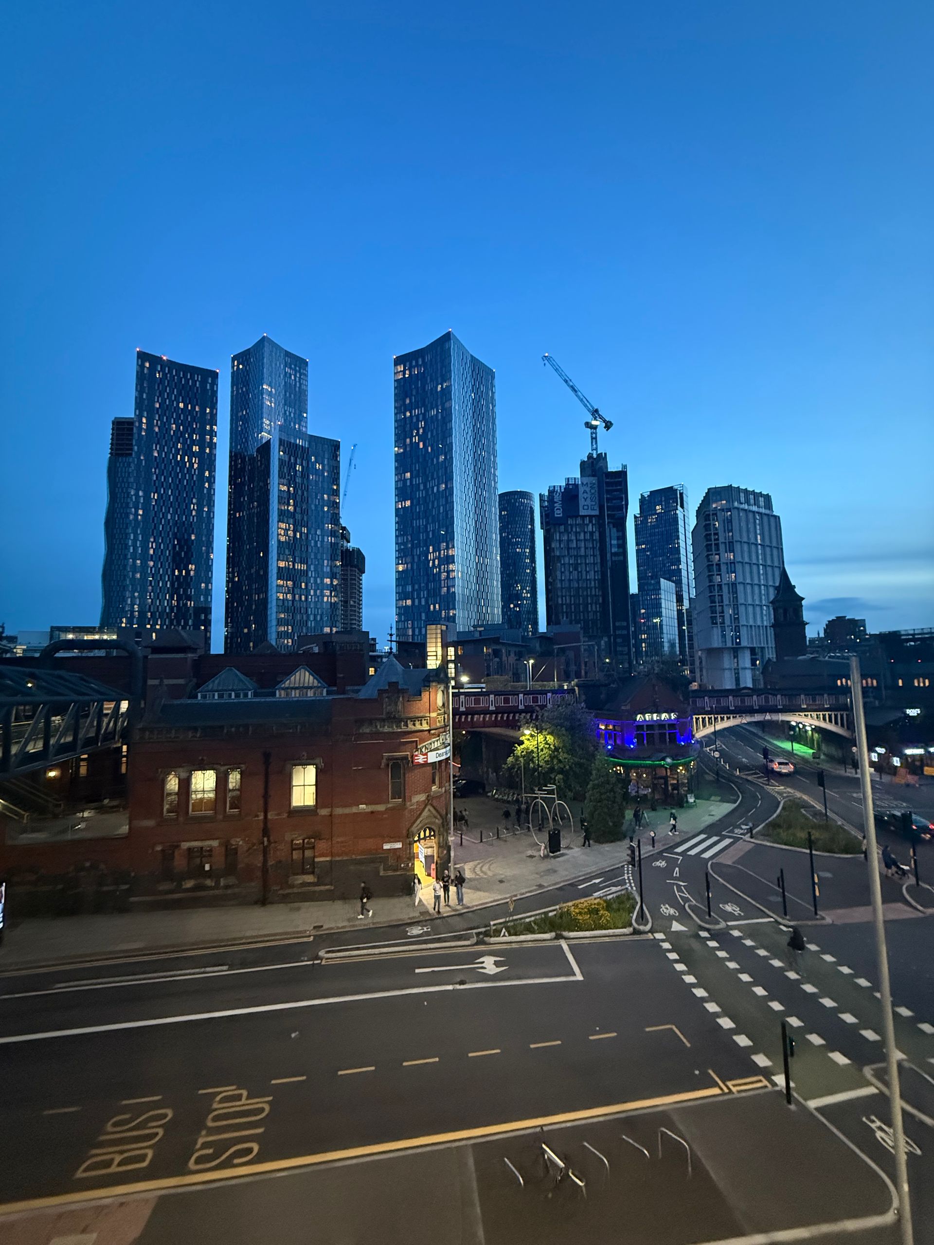 City skyline at dusk. Tall buildings with lit windows stand behind a street with a brick building and a roundabout.