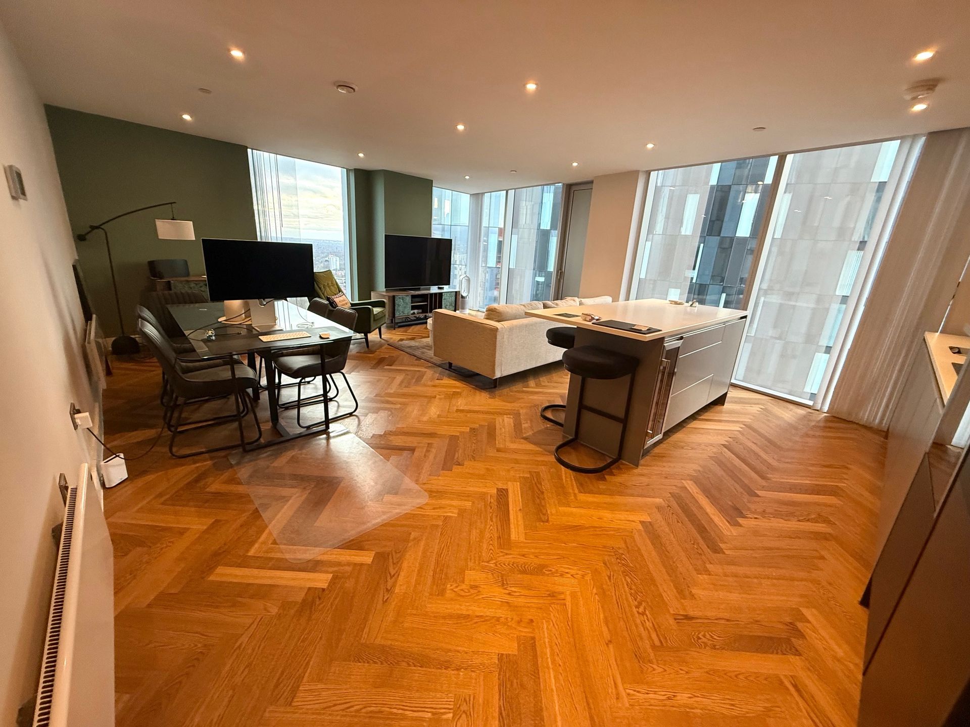 Modern living room with herringbone wood floors, green accent wall, large windows, and contemporary furniture.