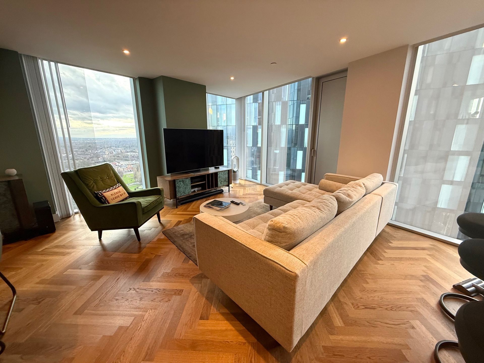 Living room with large windows, light couch, green armchair, TV, and wood floors.