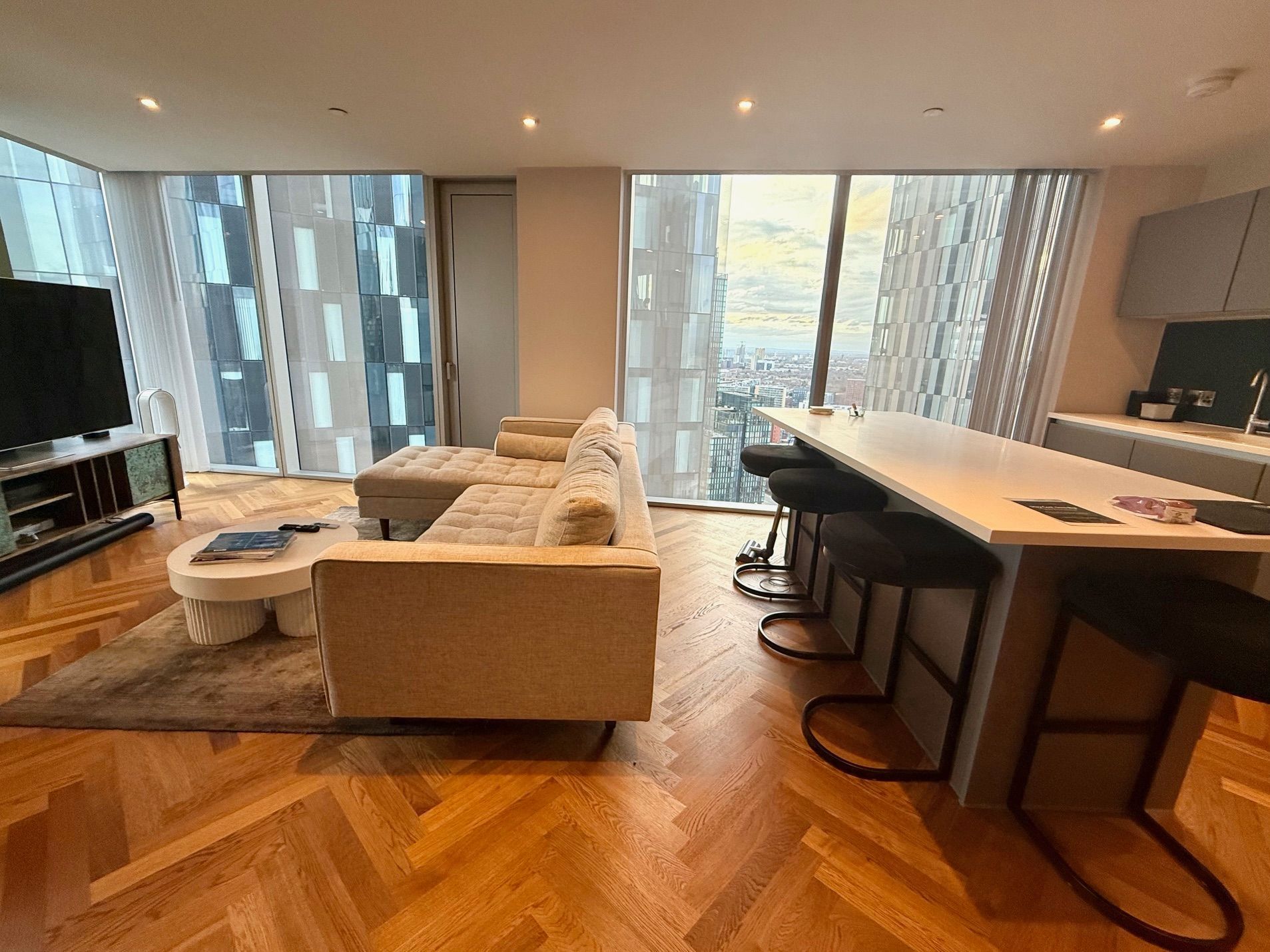 Modern living room with floor-to-ceiling windows, sofa, TV, kitchen island with stools, and wooden flooring.