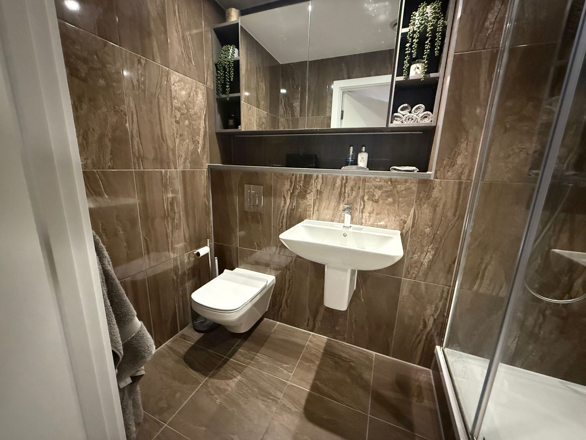Modern bathroom with brown tile, white sink, toilet, and shower. Mirror and shelves with plants are present.