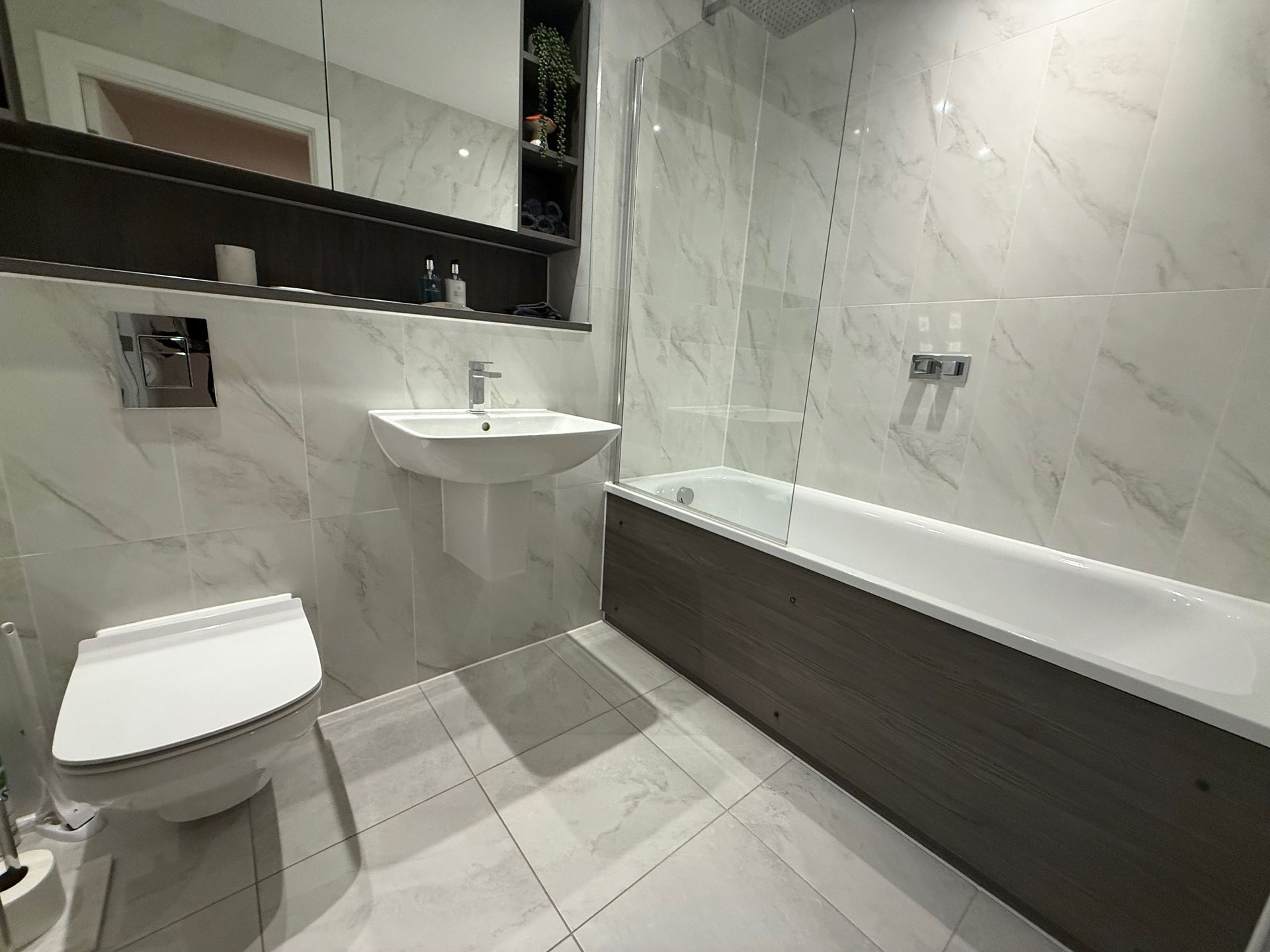 Modern bathroom with white marble-look tiles, toilet, sink, and wood-paneled bathtub.